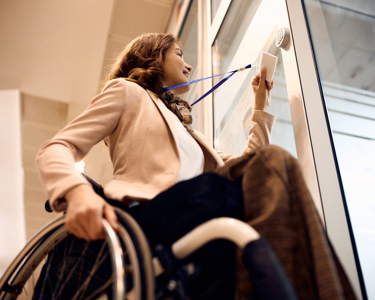 A woman using a wheelchair approaches an office door to swipe an electronic entry badge