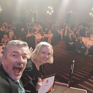 Two formally dressed professionals on stage taking a selfie at an awards banquet.