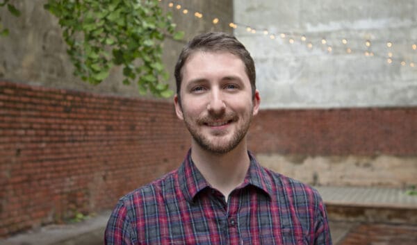 Daniel Rauh, a young man with man hair, mustache, and beard, smiles at the camera