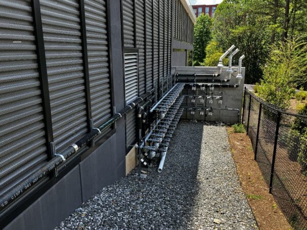 On the back side of a large building three large horizontal pipe and several smaller pipes lead to a row of gauges and meters