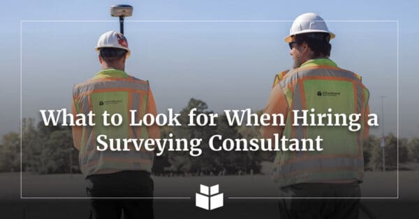 Two surveyors wearing hard hats and safety vests conduct fieldwork with GPS surveying equipment, with the text “What to Look for When Hiring a Surveying Consultant” overlaid on the image.
