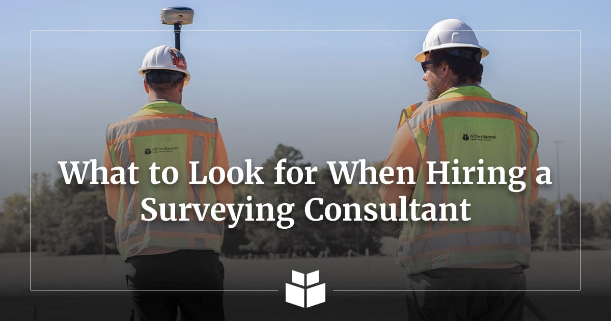 Two surveyors wearing hard hats and safety vests conduct fieldwork with GPS surveying equipment, with the text “What to Look for When Hiring a Surveying Consultant” overlaid on the image.