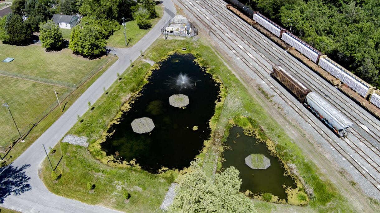 Aerial view of two stormwater ponds with vegetated islands, a fountain, and a fenced pump station beside railroad tracks.