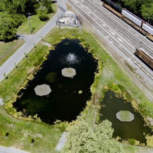 Aerial view of two stormwater ponds with vegetated islands, a fountain, and a fenced pump station beside railroad tracks.